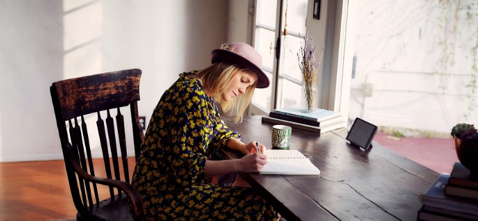 vrouw met hoed zit aan tafel te schrijven met pen