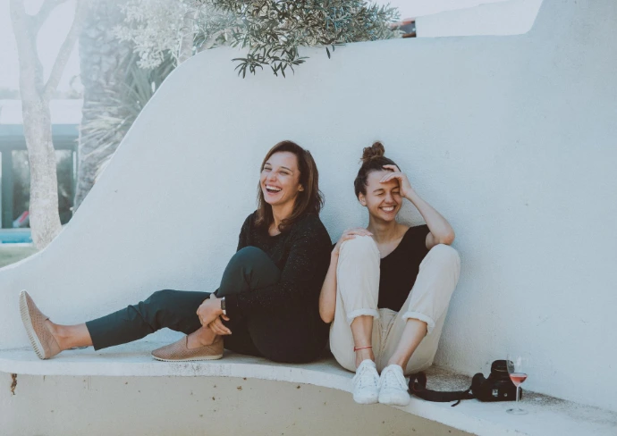twee lachende jonge vrouwen zitten samen op grond tegen muur